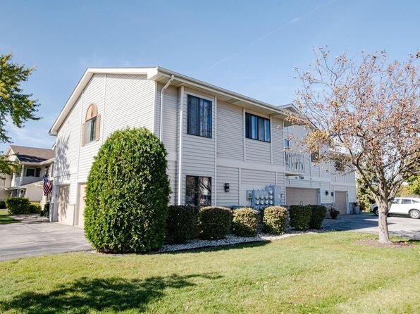 A light-colored, two-story residential building with multiple units, featuring a well-maintained front yard. A tall bush on one side and a tree with sparse autumn leaves add to the neat landscaping, while the driveway provides access to multiple garages.