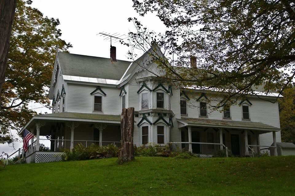 A large Victorian-style home with a white exterior, featuring intricate green trim around the windows and roof edges. The house sits atop a grassy hill with trees in the background, giving it a rustic, historical appearance.