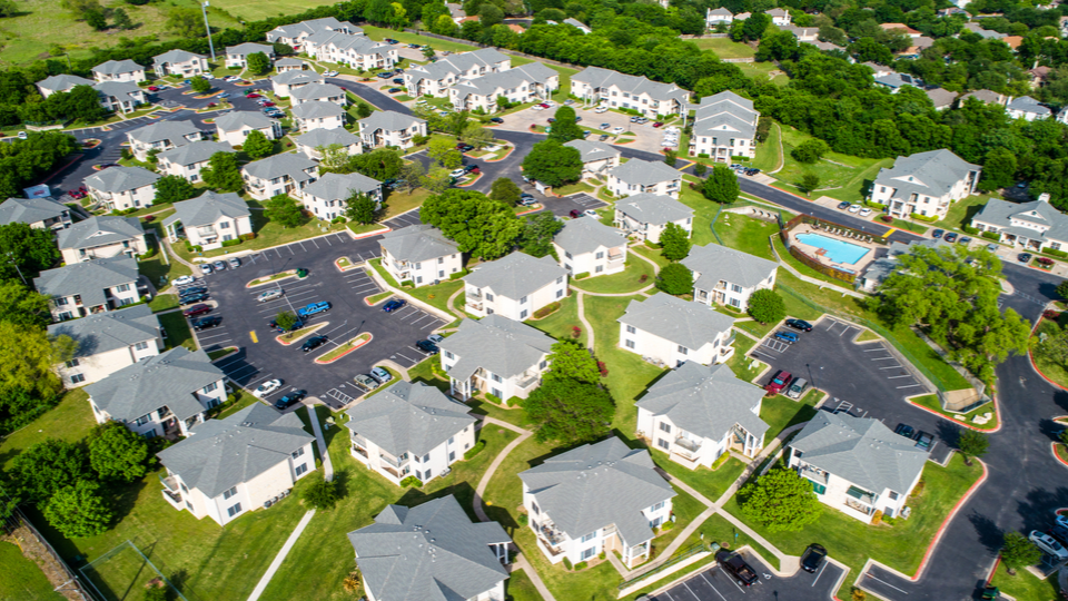 A bird's-eye view of a suburban neighborhood featuring rows of neatly organized, identical houses with grey rooftops. The area includes green lawns, a community pool, and parking spaces, showcasing a well-planned residential complex surrounded by trees.