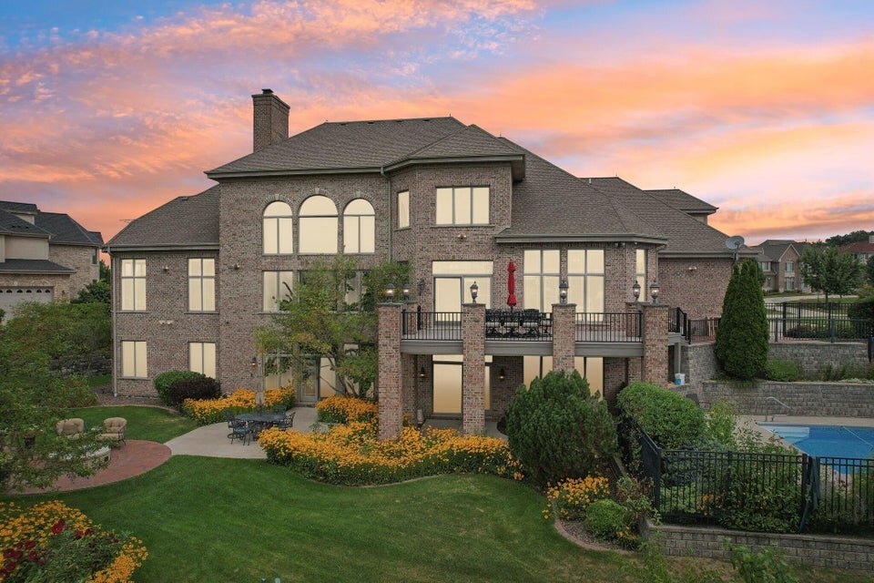 A grand two-story brick home with large arched windows and a spacious balcony, set against a beautiful sunset sky. The backyard features well-kept landscaping with blooming flowers, a paved patio area for seating, and a black iron fence enclosing the property, giving it an elegant and private feel.