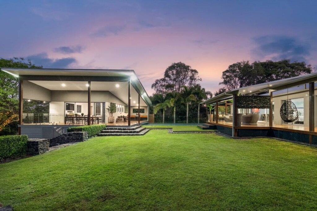 A modern, open-concept home with extensive glass walls, surrounded by lush greenery and lit with outdoor lights in the evening. The home features a spacious outdoor seating area, a large, manicured lawn, and scenic landscaping, creating a relaxing atmosphere under a twilight sky.