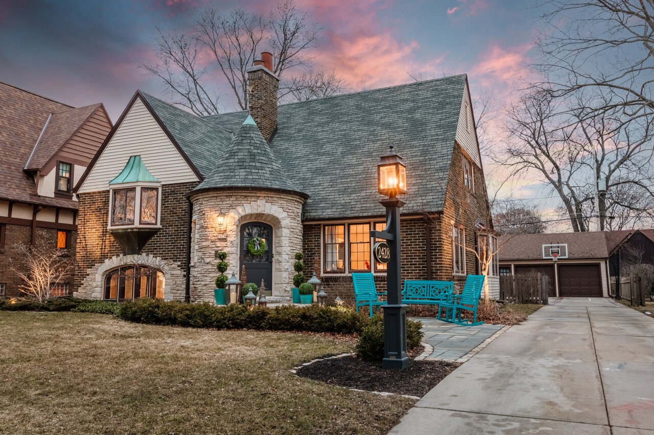 A Tudor-style home with stone and brick detailing, featuring a rounded front turret and a front door framed by an arched stone entryway. The house is lit warmly against a colorful sunset sky, creating a cozy and inviting appearance, with a pathway leading to the entrance.