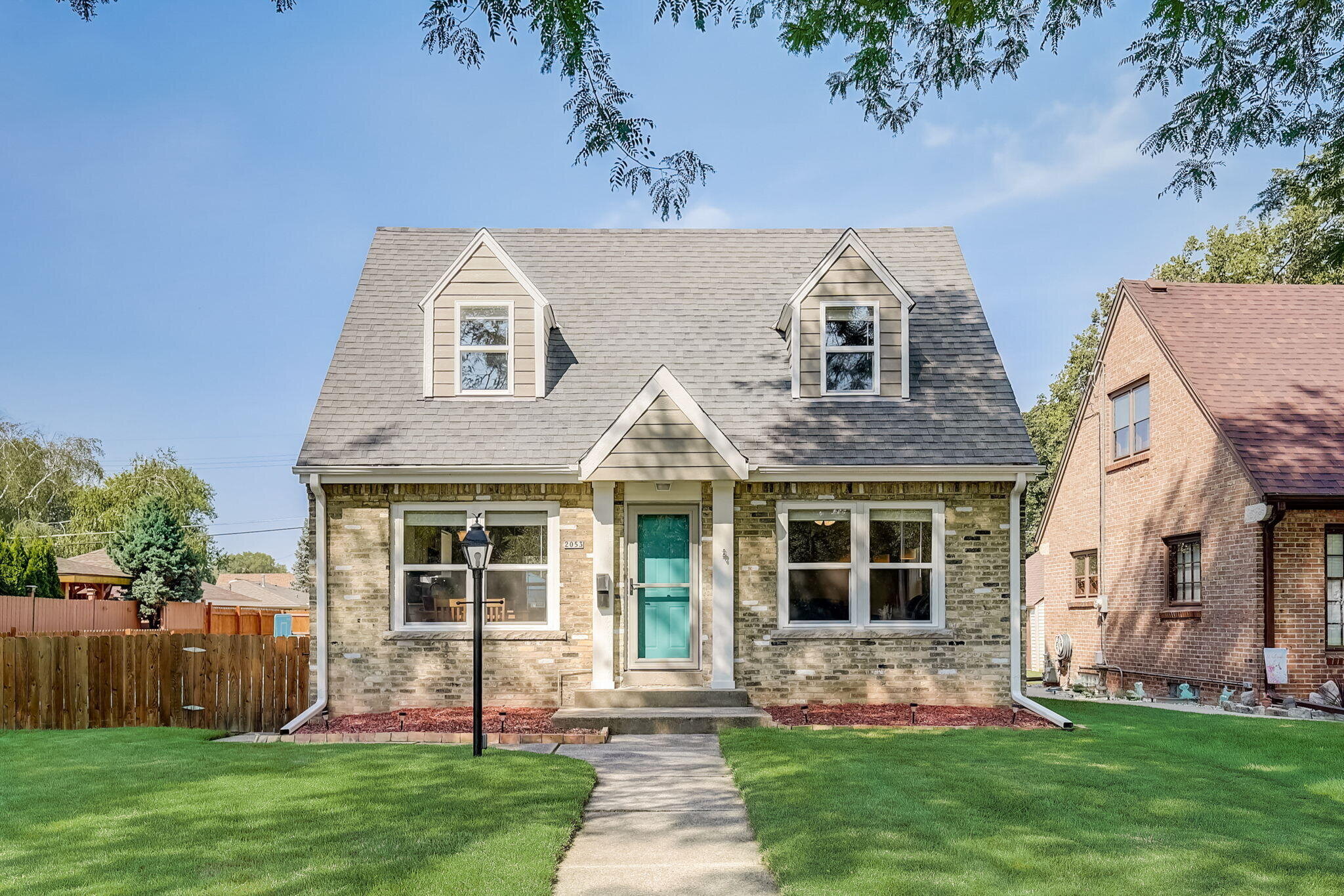 A well-maintained Cape Cod-style home with a symmetrical facade, a steep roof with dormer windows, and a bright teal front door. It has a small lawn, a paved path, and is nestled between other brick houses.