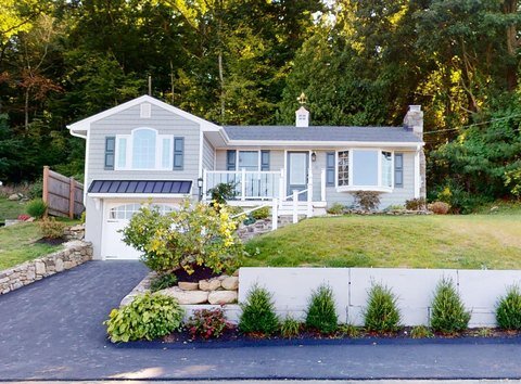 A charming, light grey split-level house with a black metal awning over the front windows. The home is set against a lush, wooded background with a driveway leading up to it. The front yard is elevated, with stone steps and manicured bushes lining the path to the entrance.