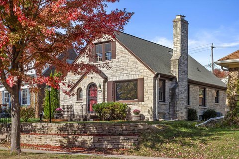 A charming stone cottage-style home with an arched red front door, surrounded by vibrant autumn foliage. The house features a sloping roof, a prominent chimney, and a landscaped front yard, creating a cozy, inviting appearance.
