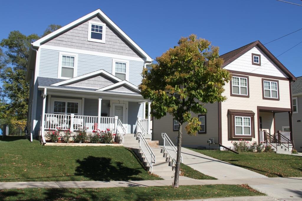 A pair of modern townhouses, one with light blue siding and the other beige, each featuring a cozy front porch, decorative trim, and a small tree out front. The manicured lawns and steps up to the doorways add a touch of suburban appeal.
