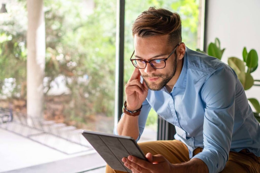 A young professional man with glasses, dressed in a light blue shirt, attentively looks at a tablet screen while seated by a large window. The lush greenery outside provides a serene backdrop, creating a relaxed and focused atmosphere as he reviews or researches information on his device.