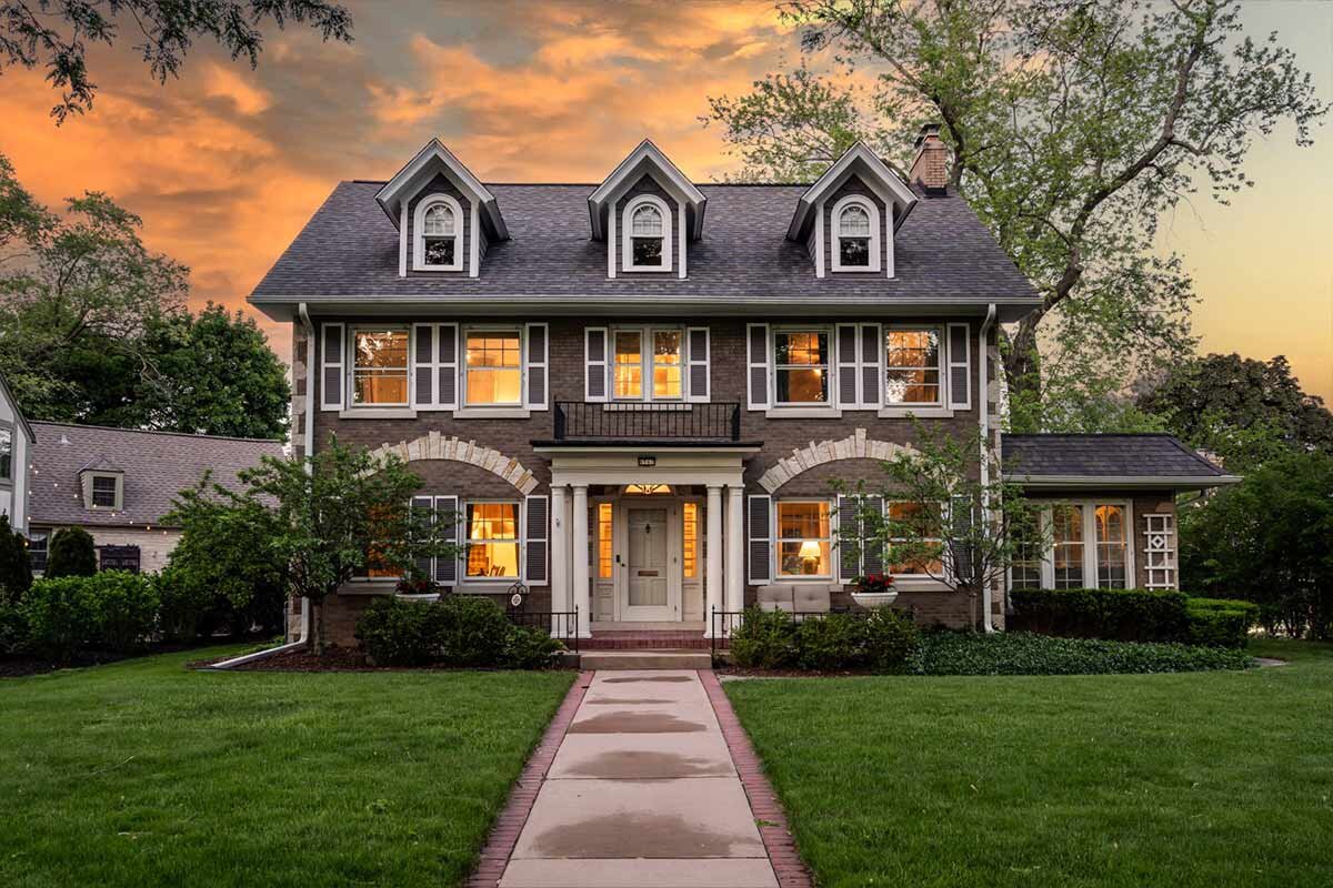 A beautiful, classic colonial-style home with symmetrical design, large front windows, and a black roof. The house is illuminated against a dramatic sunset sky, creating an inviting and picturesque scene.