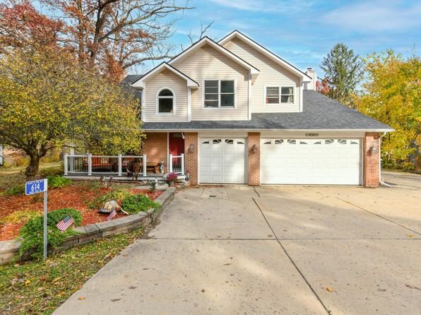 A modern two-story house with a light beige exterior and a prominent red front door, flanked by two large garage doors. The driveway is expansive, with a landscaped front yard featuring autumnal trees and an American flag.