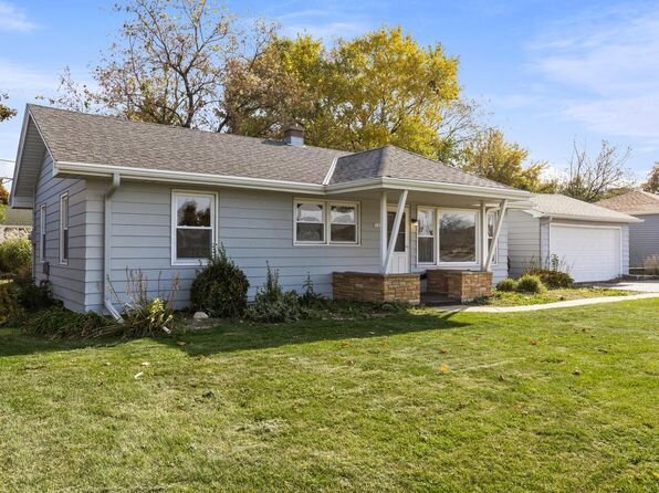 A modest single-story ranch home with light gray siding, a wide front lawn, and mature trees in the background. The house features a simple, functional design with large windows and a small front porch area.