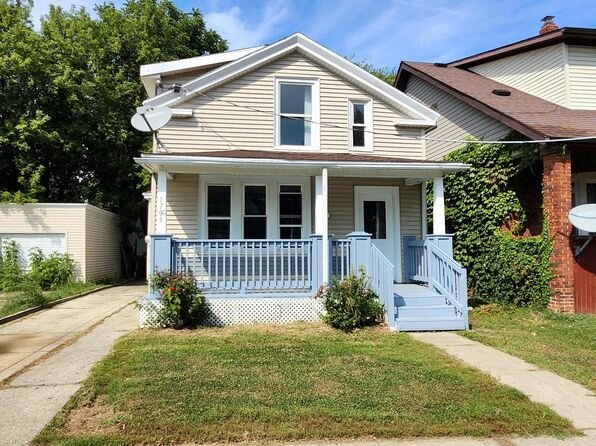 A quaint beige house with blue accents and a welcoming front porch. It sits on a small lot with a simple lawn, and the house has a traditional style with two satellite dishes attached to the exterior.