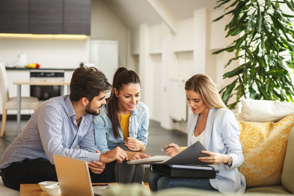A real estate agent sits on a comfortable couch, showing documents in a binder to a young couple who appear engaged and interested. The couple leans forward, listening and discussing the details, suggesting they are considering a property or financial options. The setting is a bright, modern living room with stylish decor, including a large green plant and yellow-accented cushions, creating a warm and welcoming atmosphere.