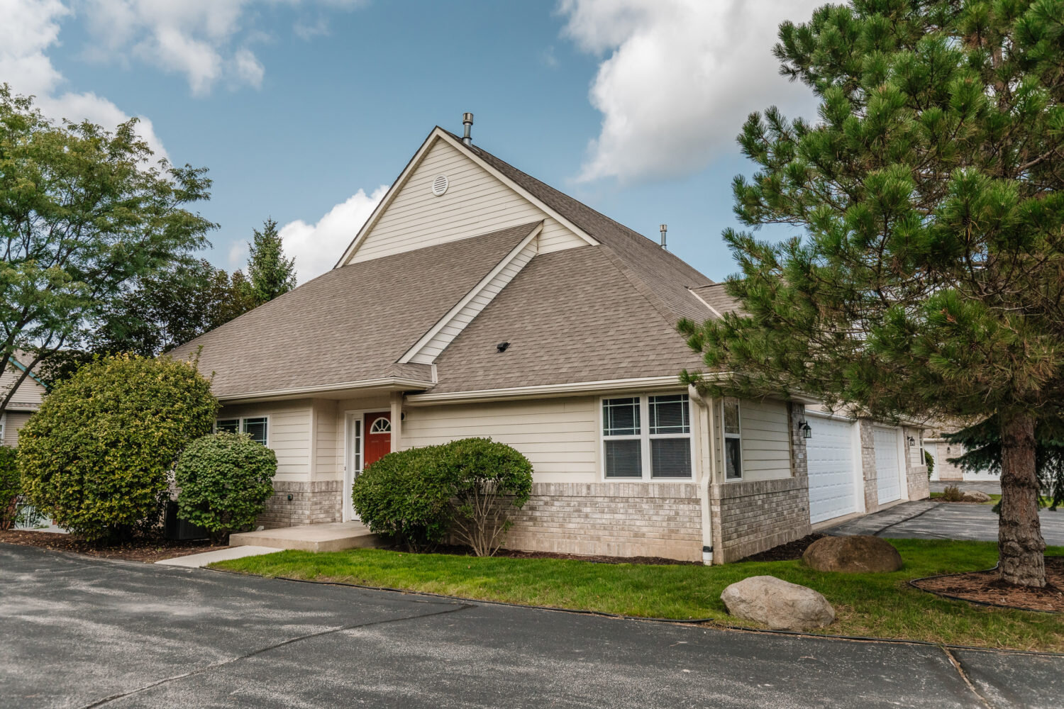 A single-story, ranch-style home with a spacious driveway and a well-kept lawn. Surrounded by mature trees, the house features beige siding and a welcoming red front door.