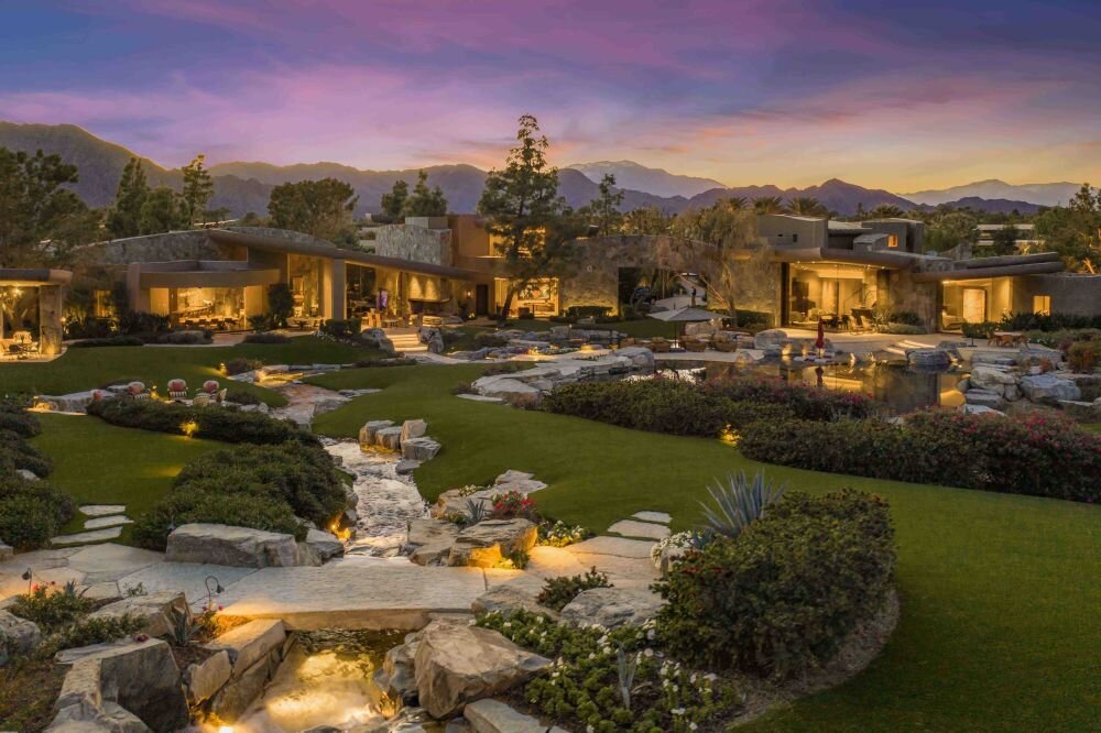 A sprawling luxury estate with modern architecture, nestled in a beautifully landscaped garden. A flowing water feature winds through the property, illuminated with soft lights as the sky turns purple with the setting sun and mountains in the background.