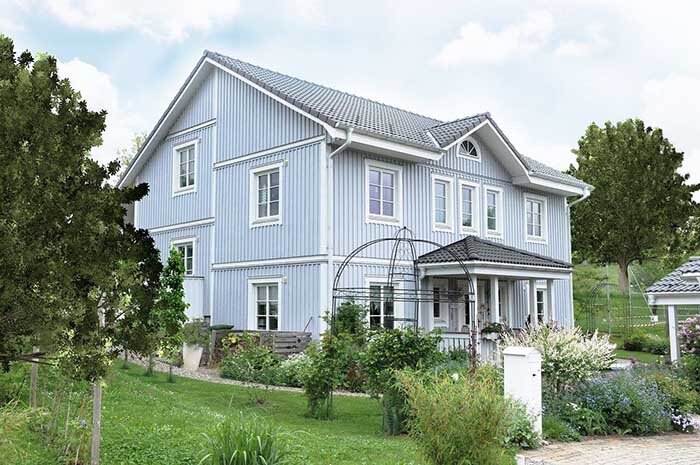 A two-story blue Scandinavian-style house with a gabled roof and large windows. The house is surrounded by a vibrant garden with various shrubs and flowering plants, offering a serene and picturesque atmosphere.