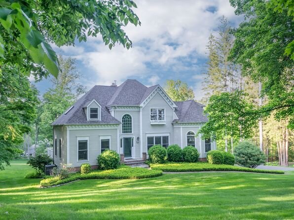 A beautiful two-story suburban home with light grey siding, tall arched windows, and a dark shingled roof, set amidst lush greenery. The front yard features a well-manicured lawn, rounded hedges, and a landscaped path leading to the entrance. Mature trees surround the property, creating a serene, natural backdrop against a partly cloudy sky.