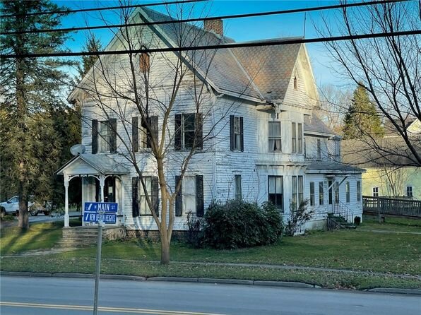 An aged, weathered white house with a steeply pitched roof and a wrap-around front porch. The building shows signs of wear but retains its historic charm, positioned on a corner lot with a "Main St" street sign in view. The yard is partially enclosed by a hedge and some bare trees, giving the house a rustic, vintage look.