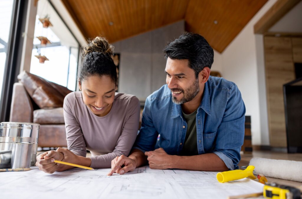 A smiling couple lies on the floor, reviewing home renovation plans together. The woman holds a pencil while the man points to details on the blueprint, both appearing engaged and enthusiastic about the project. Around them are tools, paint cans, and a paint roller, suggesting they are preparing for a home improvement or remodeling task. The warm interior and natural lighting create a cozy atmosphere, emphasizing teamwork and excitement in personalizing their living space.