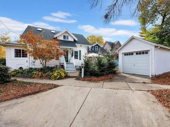A charming, white single-story home with a detached garage, surrounded by vibrant autumn trees. The house features a green roof, a quaint front porch, and a driveway adorned with fallen leaves.