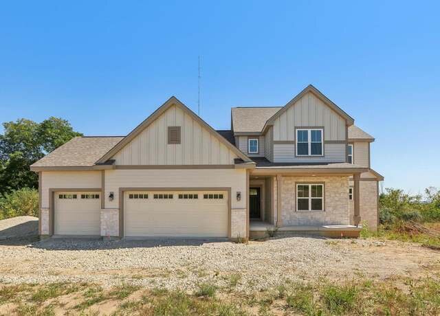  A contemporary, beige-colored house with gable roofs, large windows, and a three-car garage. The home sits on a plot of land with an unfinished yard, highlighting its recent construction and modern design.
