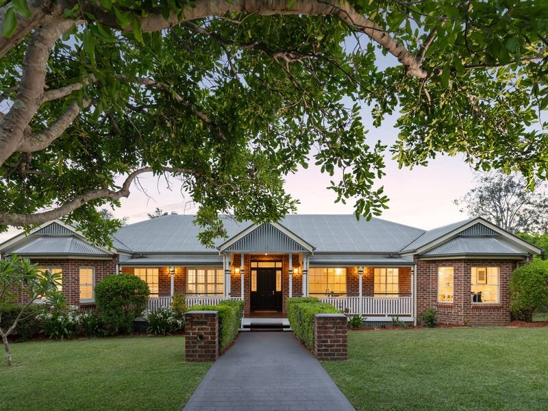 A charming brick ranch-style home with a welcoming pathway leading to the black front door, framed by white-trimmed windows and a front porch. The house is surrounded by well-maintained landscaping, including hedges and lush green grass. Overhanging tree branches create a natural frame above the property, adding a serene ambiance.