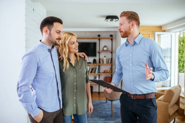 A real estate agent holding a clipboard discusses property details with a young couple inside a modern, well-lit living room. The couple, standing close together and smiling, appear attentive and engaged, suggesting a positive and friendly interaction. The room features contemporary decor, with shelves and cozy furnishings in the background.