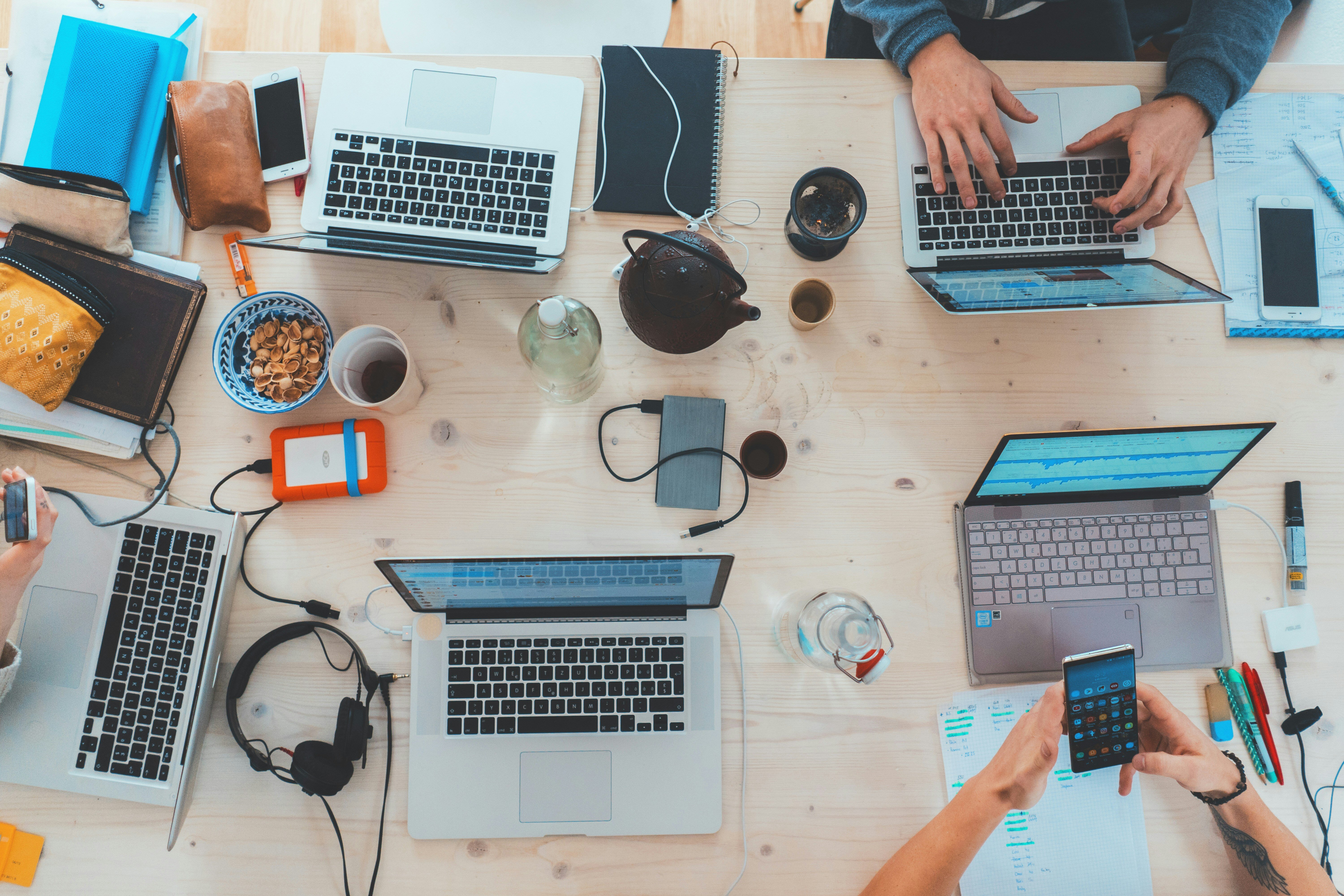 Overhead view of a collaborative workspace with multiple laptops, notebooks, and office supplies spread out on a wooden table, with individuals working on their laptops and using smartphones, creating a busy and dynamic working environment.