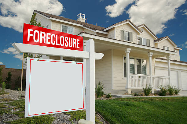 Foreclosure sign in front of a suburban house with a cloudy blue sky background.