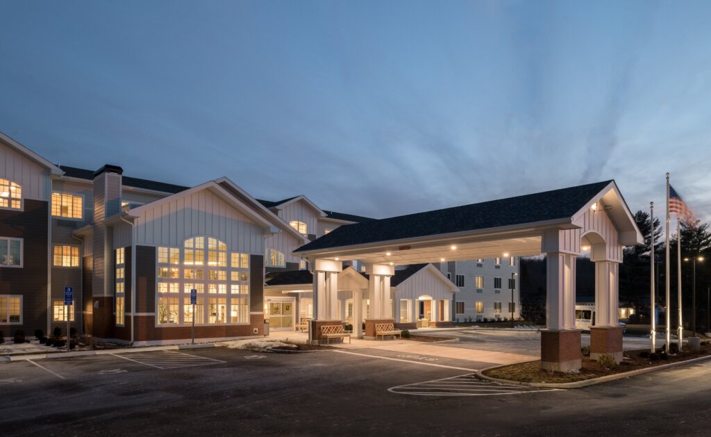 Modern residential building with large illuminated windows, a covered entryway, and an American flag under a dusk sky, surrounded by a parking lot.