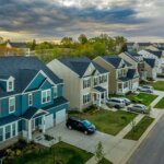 Suburban neighborhood with rows of modern two-story homes, neatly manicured lawns, parked cars, and a cloudy sunset in the background.