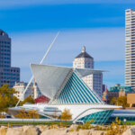 View of the Milwaukee Art Museum with its iconic, sail-like architecture against the backdrop of downtown Milwaukee. The modern building, with its striking white wings, contrasts beautifully with the surrounding skyscrapers and clear blue sky.