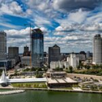 Aerial view of Milwaukee's skyline with modern buildings, a waterfront, and green spaces under a cloudy blue sky.