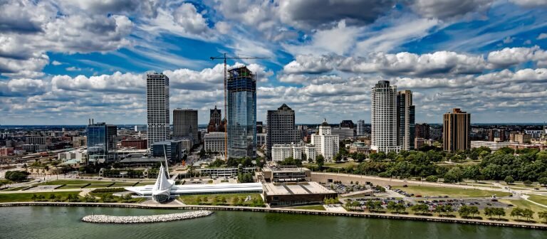 Aerial view of Milwaukee's skyline with modern buildings, a waterfront, and green spaces under a cloudy blue sky.