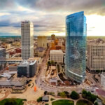 Aerial view of downtown Milwaukee showing modern skyscrapers and city streets, including the U.S. Bank building and other tall buildings. The scene is captured during sunset, with a dramatic sky in the background. The foreground shows a public park area with sculptures and green spaces, adding vibrancy to the cityscape.