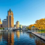 View of downtown Milwaukee along the river with autumn trees on the shore, showcasing tall buildings including a prominent skyscraper in the background, and a gazebo by the water, reflecting the clear blue sky.