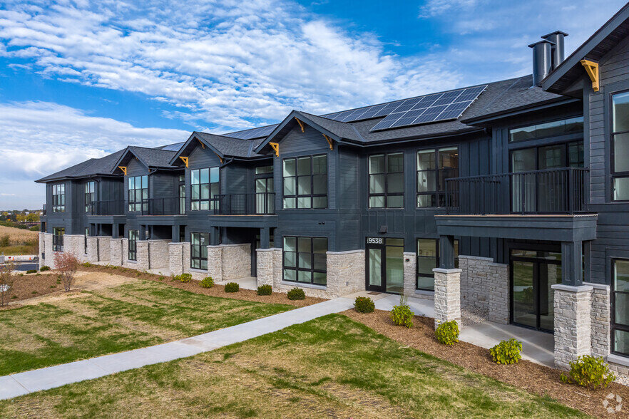 Modern apartment complex featuring dark siding, large windows, solar panels, and landscaped surroundings under a partly cloudy sky.