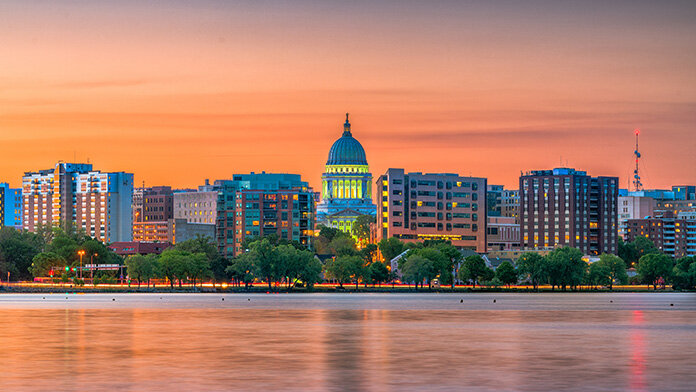 Skyline view of Madison, Wisconsin, at sunset with the illuminated Capitol building and cityscape reflected in the calm water.