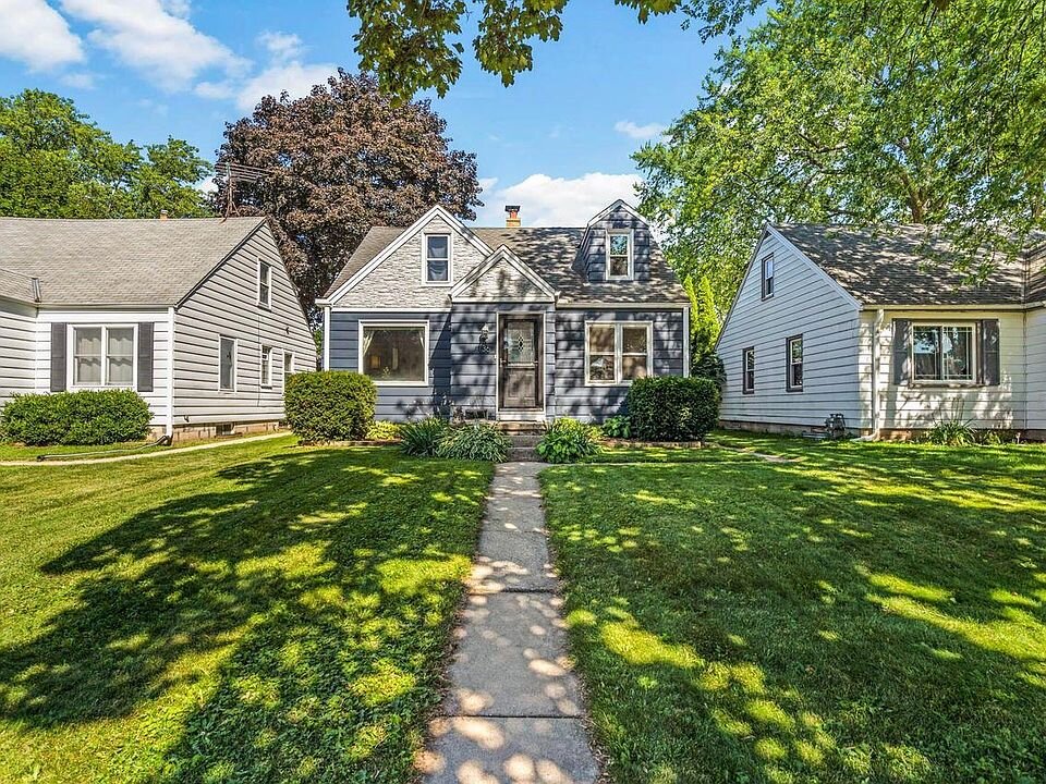 Charming suburban home with a dark gray exterior and white trim, nestled between two similar houses. A concrete walkway leads to the front entrance, surrounded by a well-manicured lawn and lush greenery under a bright blue sky.