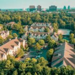 Aerial view of an apartment complex with surrounding trees and parking area, with city skyline in the distance.