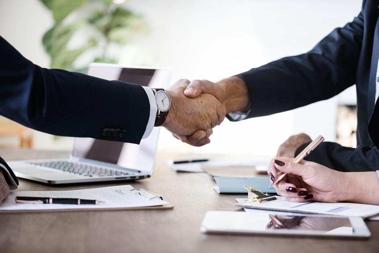 Two business professionals shaking hands over a contract, with a woman taking notes and a laptop on the table, symbolizing a successful real estate transaction.