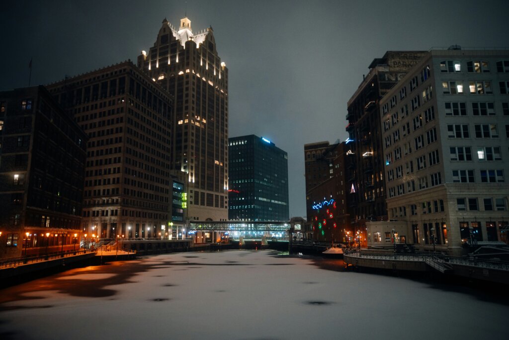 A nighttime view of Milwaukee's downtown, with tall buildings lit up and a frozen canal in the foreground, featuring colorful holiday lights on the buildings.