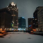 A nighttime view of Milwaukee's downtown, with tall buildings lit up and a frozen canal in the foreground, featuring colorful holiday lights on the buildings.