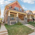 A charming two-story house with a red and beige exterior, a covered front porch, and a small front yard, situated in a row of similar residential homes.