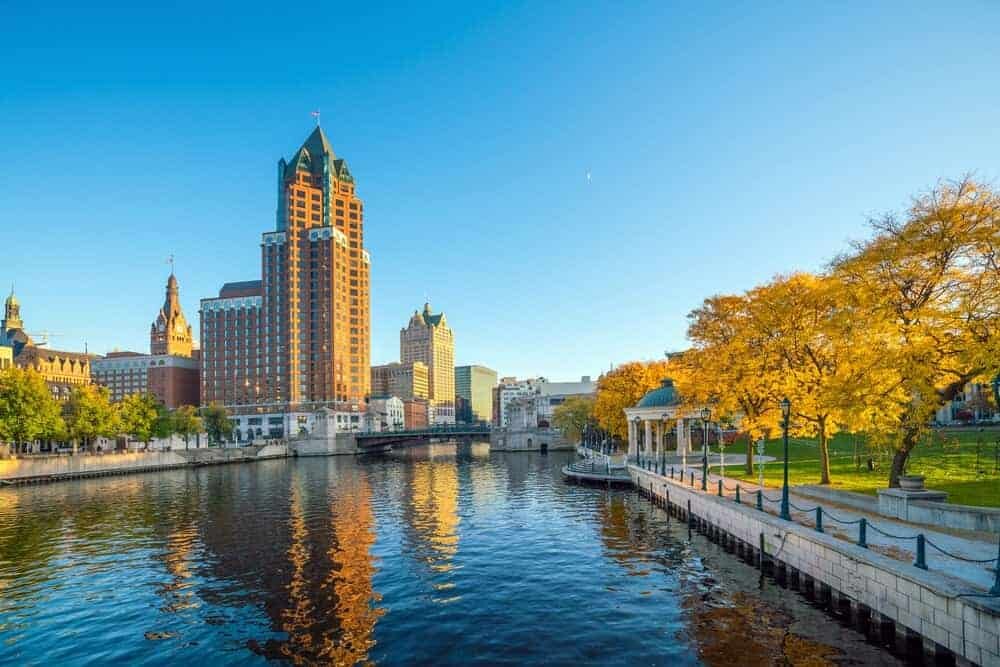 View of downtown Milwaukee along the river, featuring modern skyscrapers, autumn trees, and a scenic park with a gazebo.