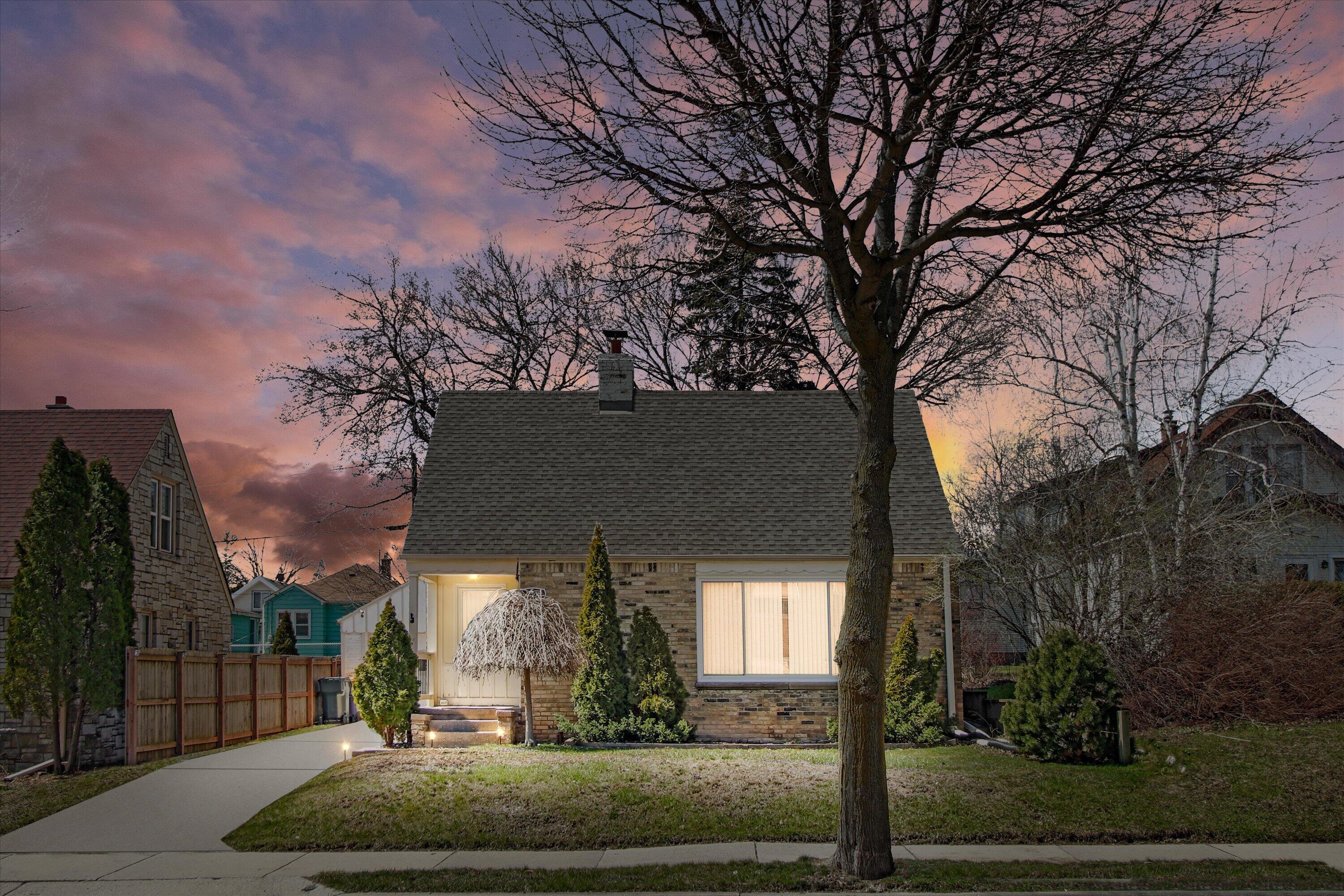 A warmly lit brick house with a gabled roof under a colorful sunset sky, surrounded by trees and a well-kept yard.