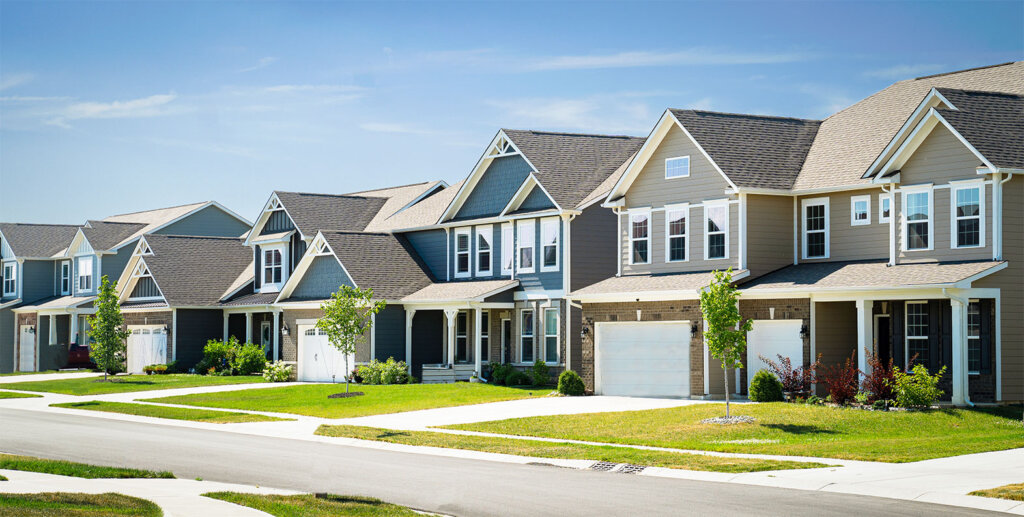 A row of modern suburban houses with gabled roofs, manicured lawns, and driveways along a quiet residential street under a clear blue sky.