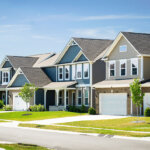 A row of modern suburban houses with gabled roofs, manicured lawns, and driveways along a quiet residential street under a clear blue sky.