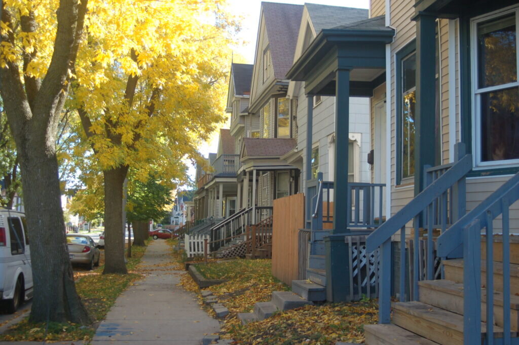 A row of traditional wooden houses with front porches in a Milwaukee neighborhood, lined with trees showcasing vibrant yellow autumn leaves.