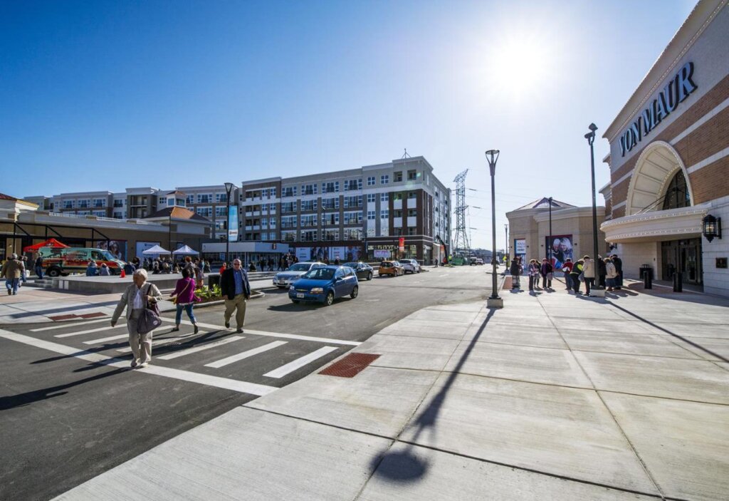 A busy shopping district with people crossing the street near Von Maur on a sunny day. Modern buildings, parked cars, and vendor tents add to the lively scene.