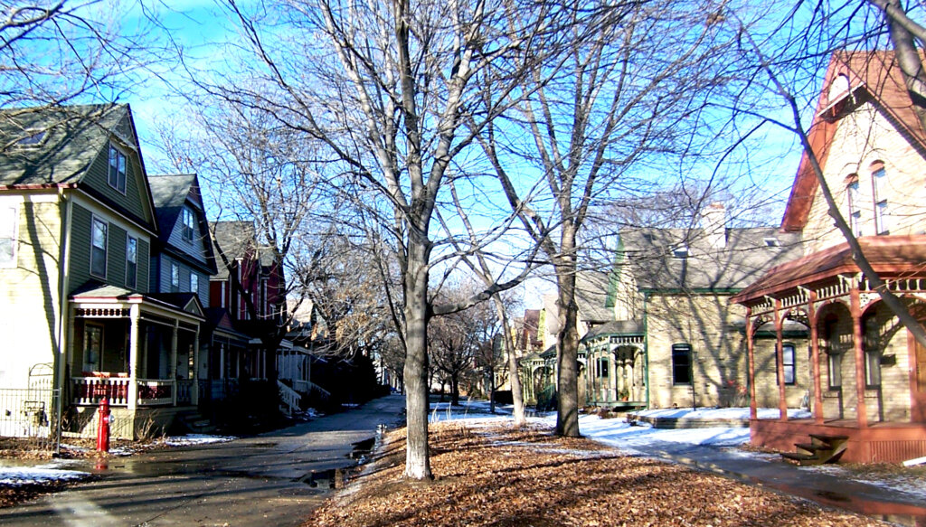 A residential street in Milwaukee featuring houses with various architectural styles, bare trees, and scattered leaves on the ground.